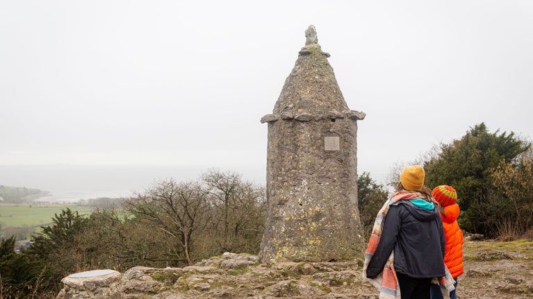 Two children in coats and wooly hats looking up at the Pepperpot monument, a large stone monument with a pointed top, at Arnside and Silverdale.